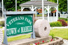 Veterans Park Town of Markle sign in white text on a green background with a white gazebo five flagpoles in the background.