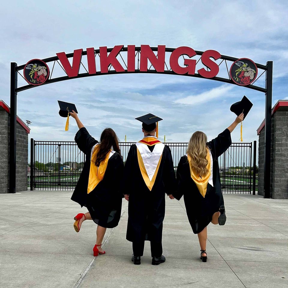 Three high school graduates in graduation gowns celebrating under the Vikings sign.