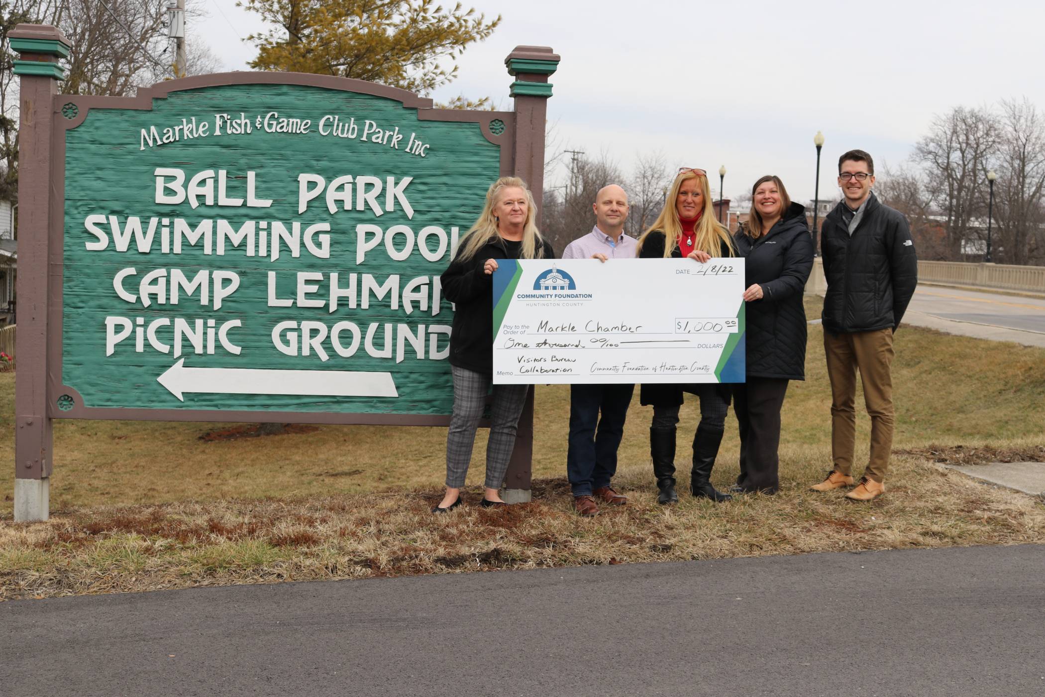 Five people holding a donation check from the Markle Fish and Game Club park in front of their sign