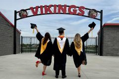 Three recent graduates wearing gowns celebrating under the Vikings sign.